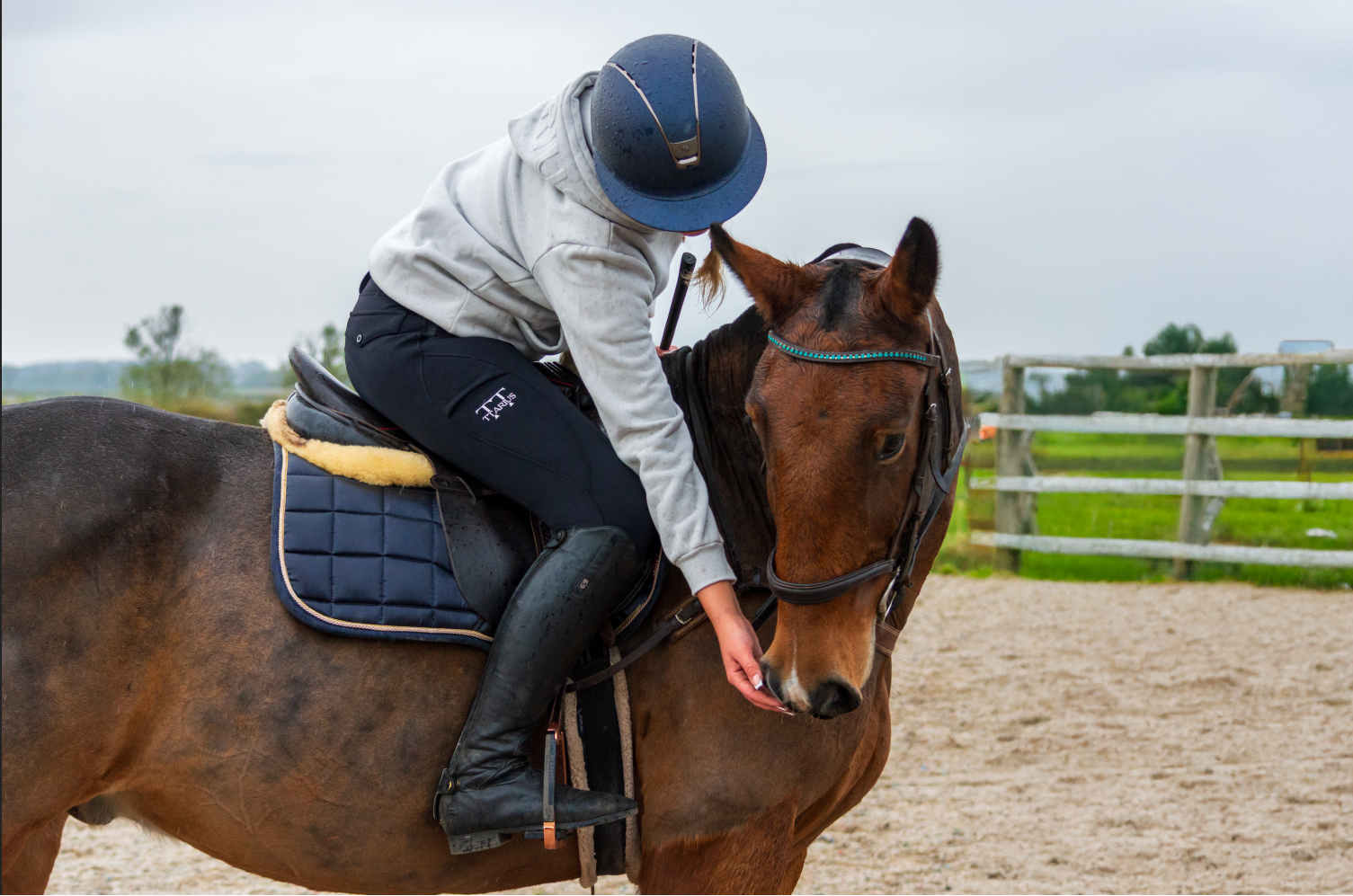 Person riding a horse in an outdoor setting with a fence and grass in the background wearing ttarius equestrian sports clothing.